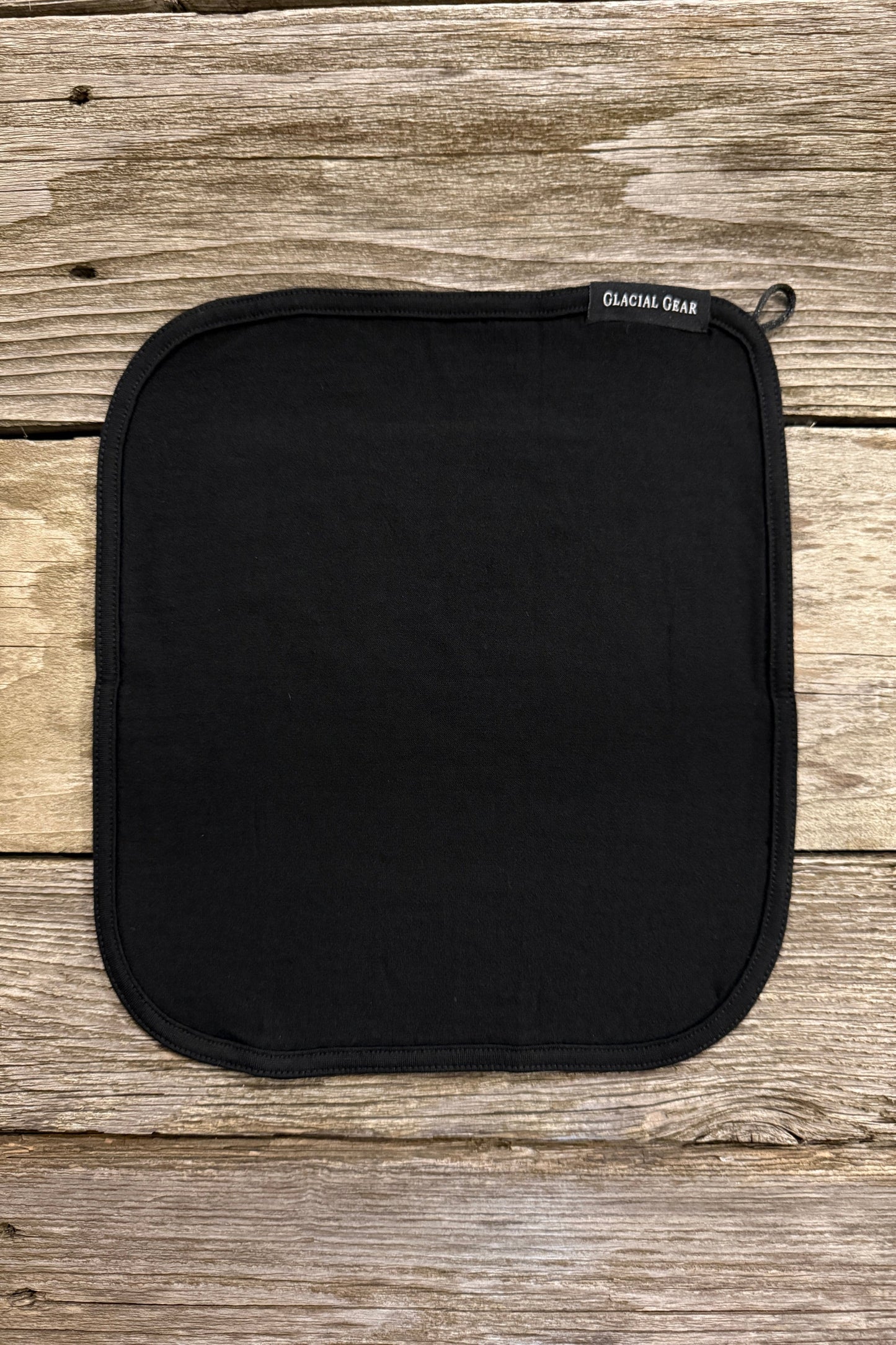 A black merino wool trail towel with a corner clip, laid flat on a wooden surface.