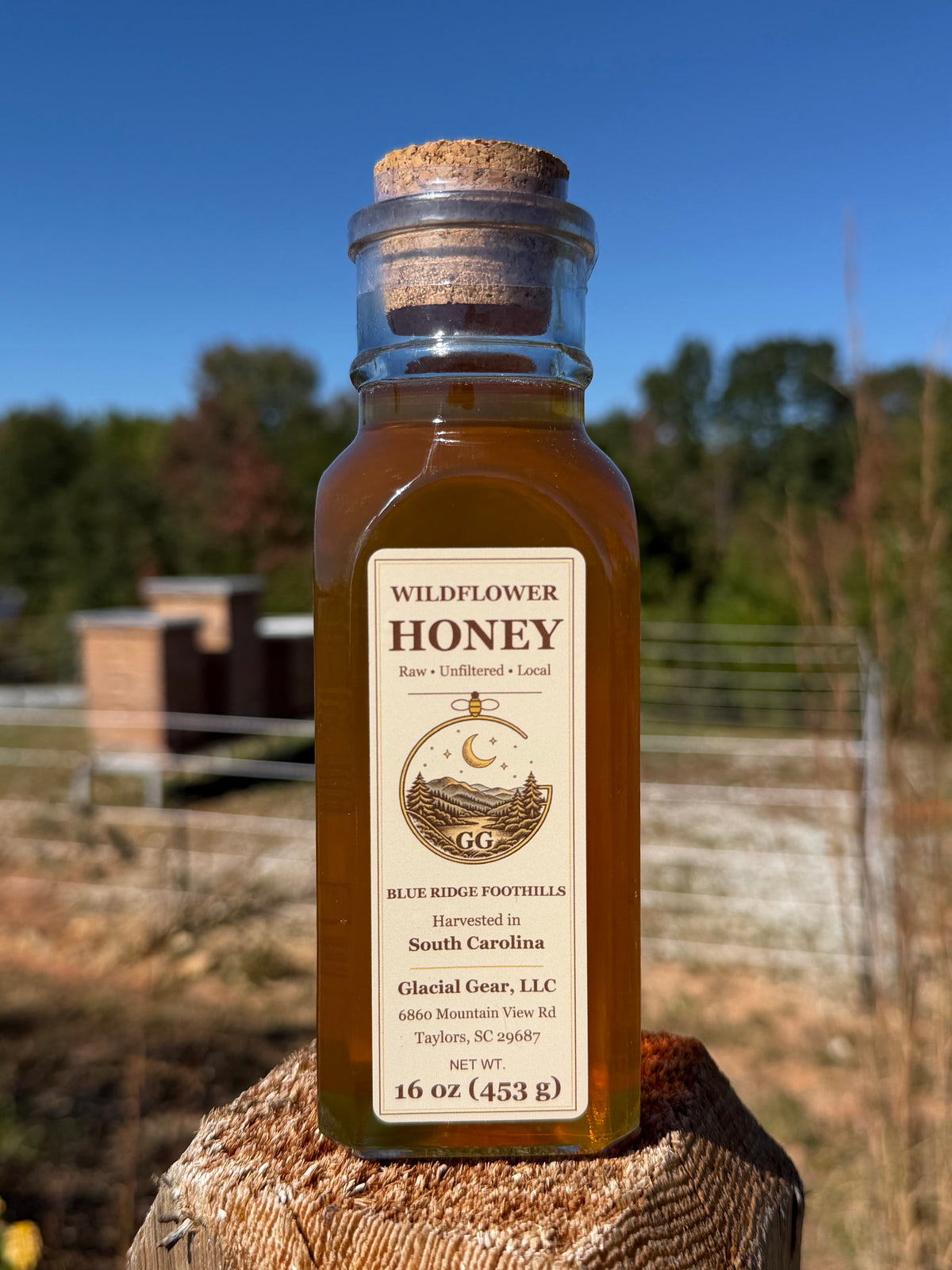 GG Apiary wildflower honey in a corked Muth glass jar on a wooden post, with our beehives behind the fence.