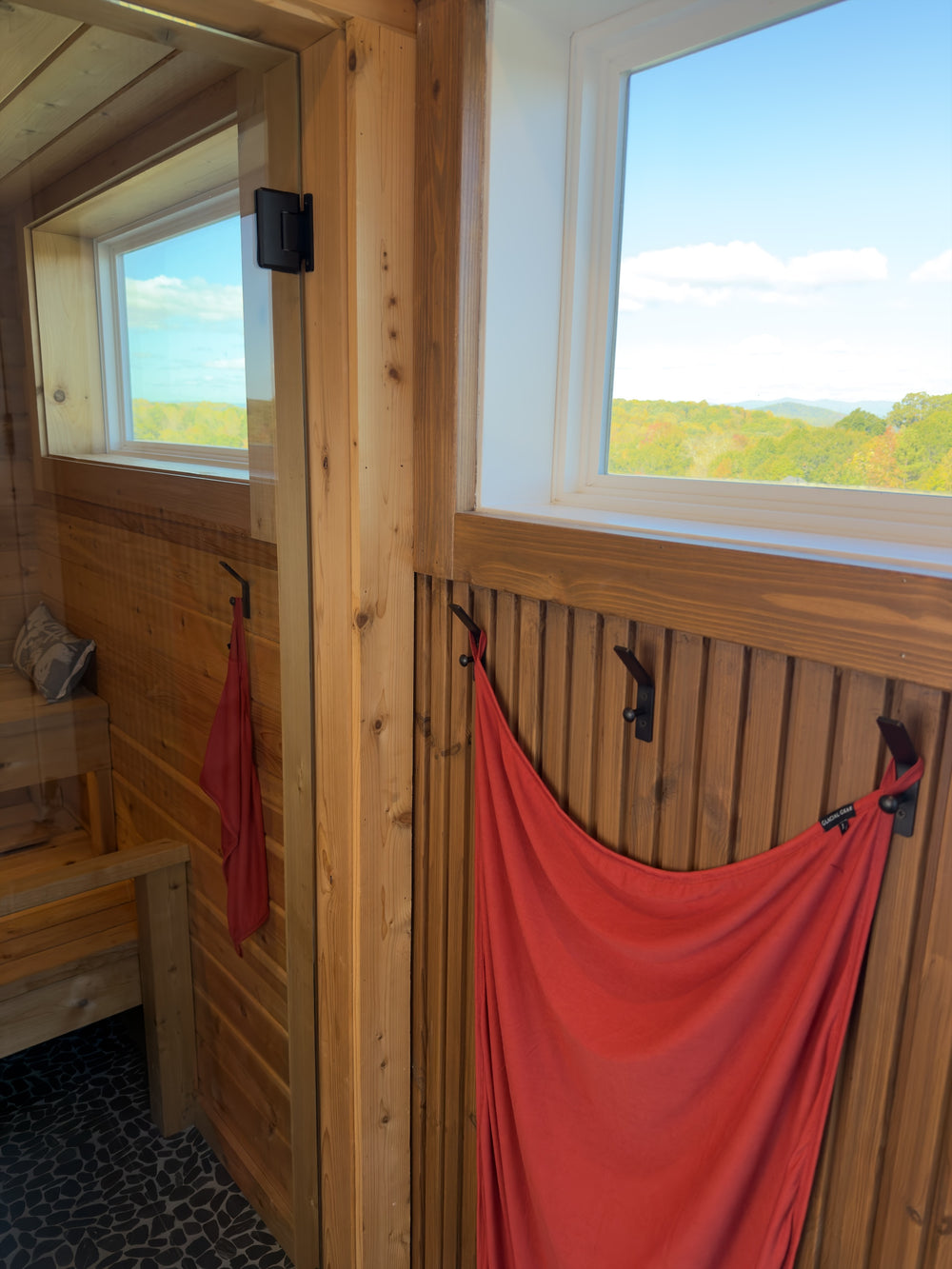 Autumn Glacial Gear x Zpacks Camp Towels hanging by their corner loops inside and outside a sauna, with a window view of fall trees and mountains