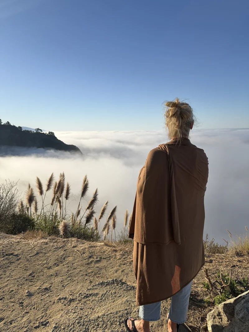 Shereen wrapped in a brown merino blanket, with warm sunlight on her shoulders, overlooking clouds and a wooded ridge