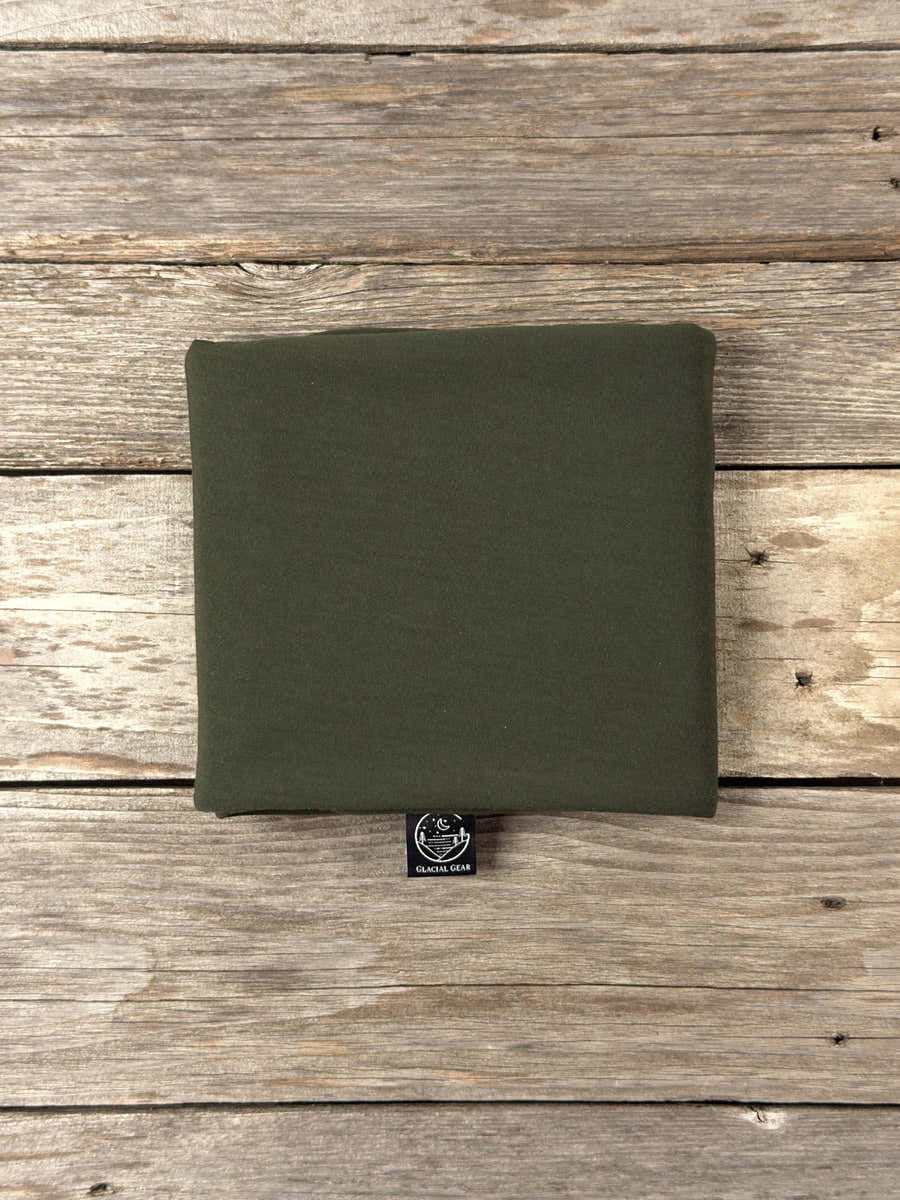 Folded forest green Glacial Gear Merino wool sleeping bag liner with logo tag on weathered wood