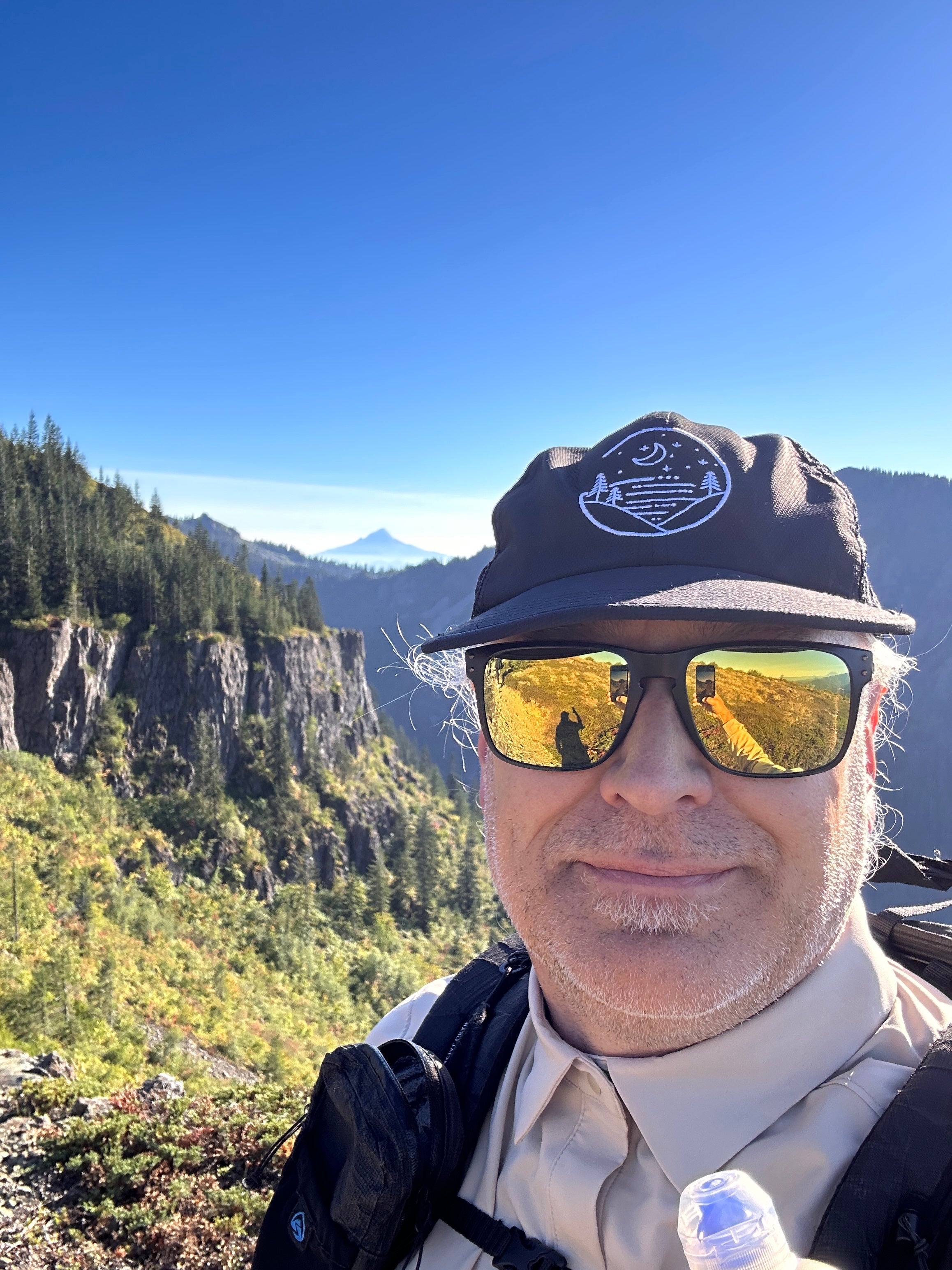 Shane wearing a black Glacial Gear Foldable Trail Hat and sunglasses above forested mountain cliffs.