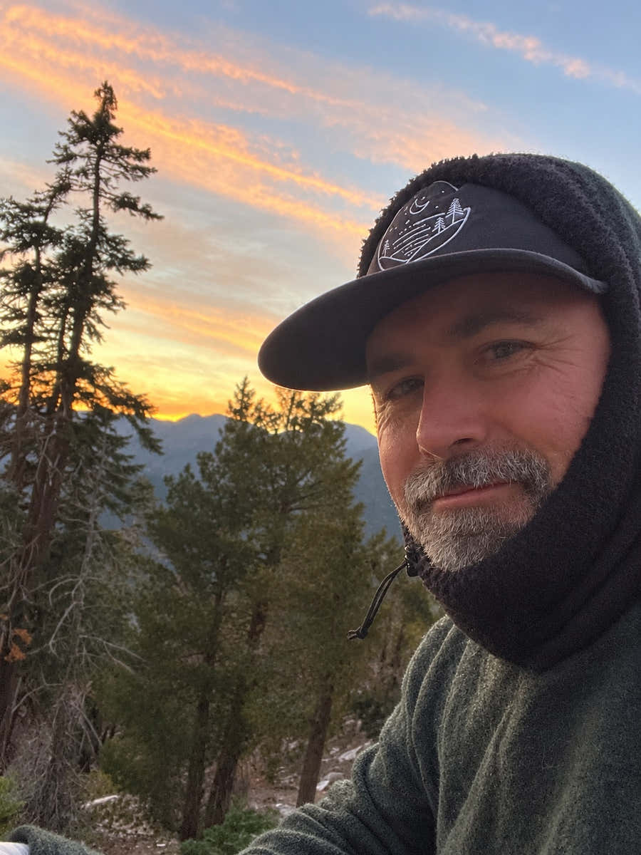Hiker wearing a black Glacial Gear Foldable Trail Hat beneath a hood, with evergreen trees and mountain ridges at sunset.