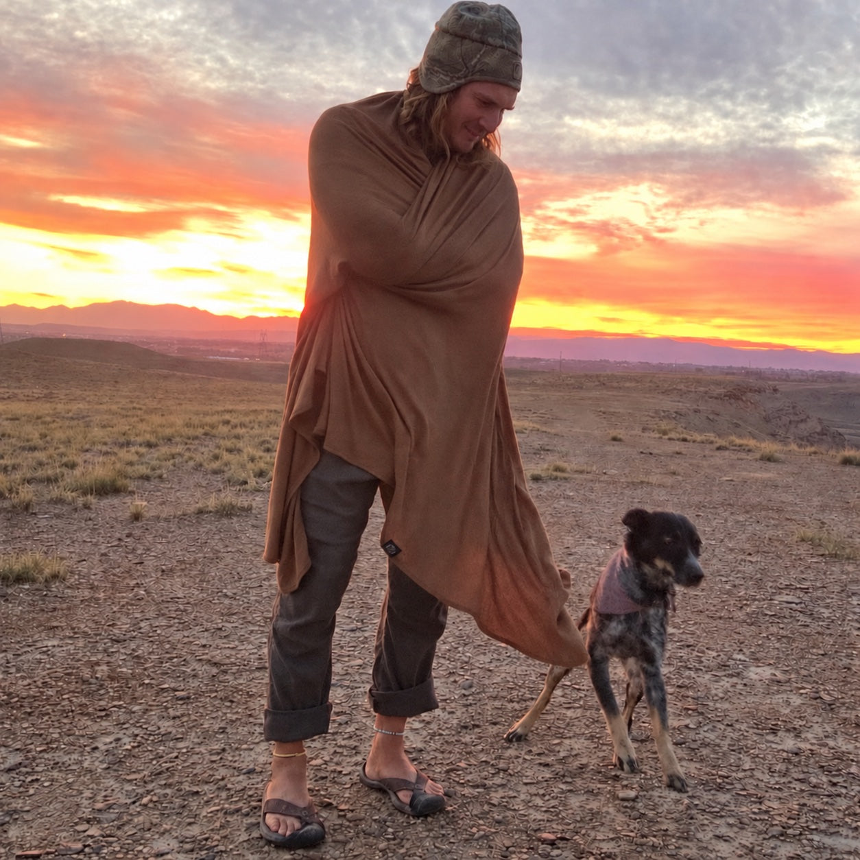 Hiker wrapped in a Bark brown Glacial Gear ultralight Merino wool blanket beside a dog at sunset.