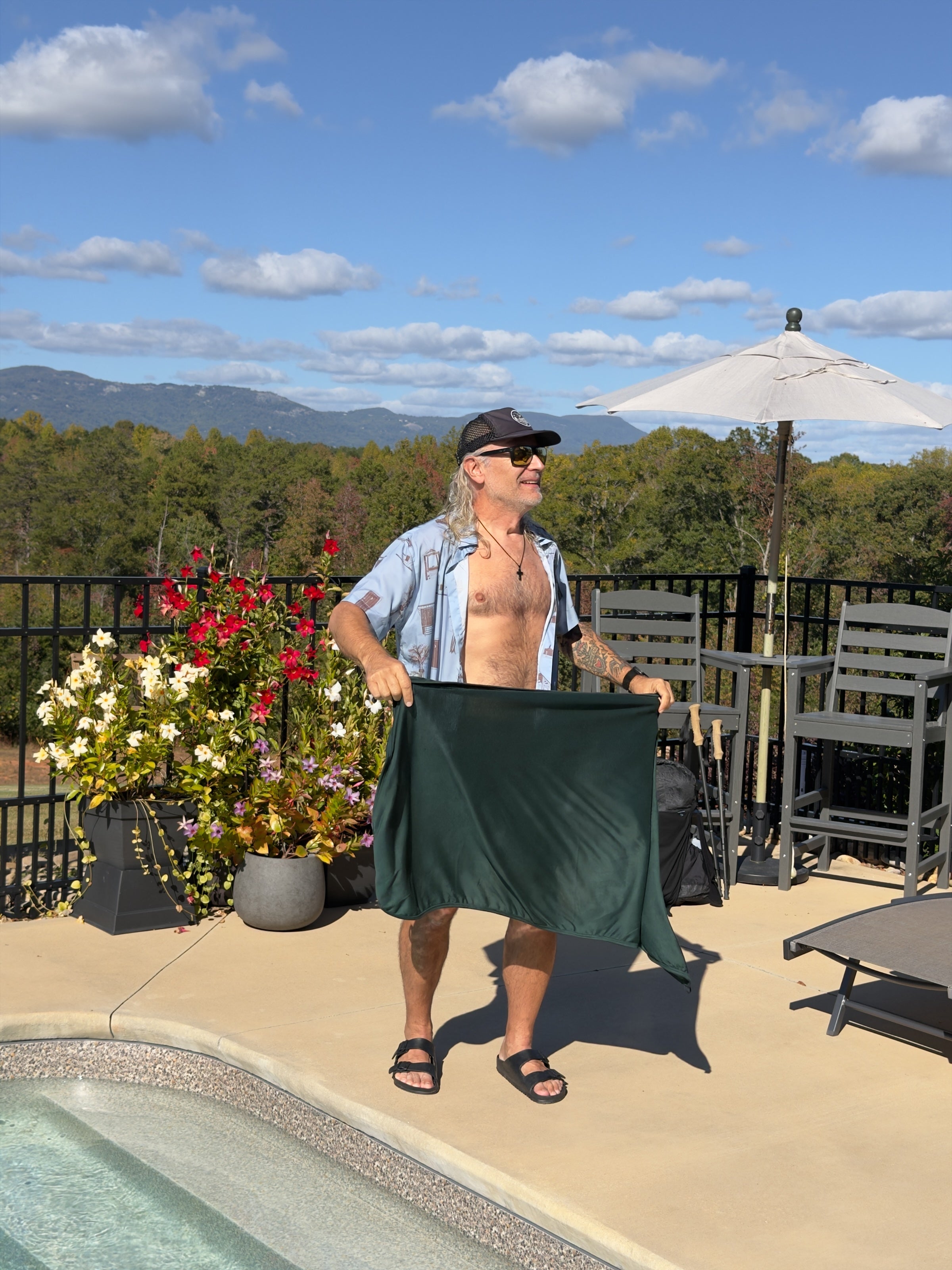 Shane holding a Forest Glacial Gear x Zpacks Camp Towel open by a pool, with fall trees and mountains behind him