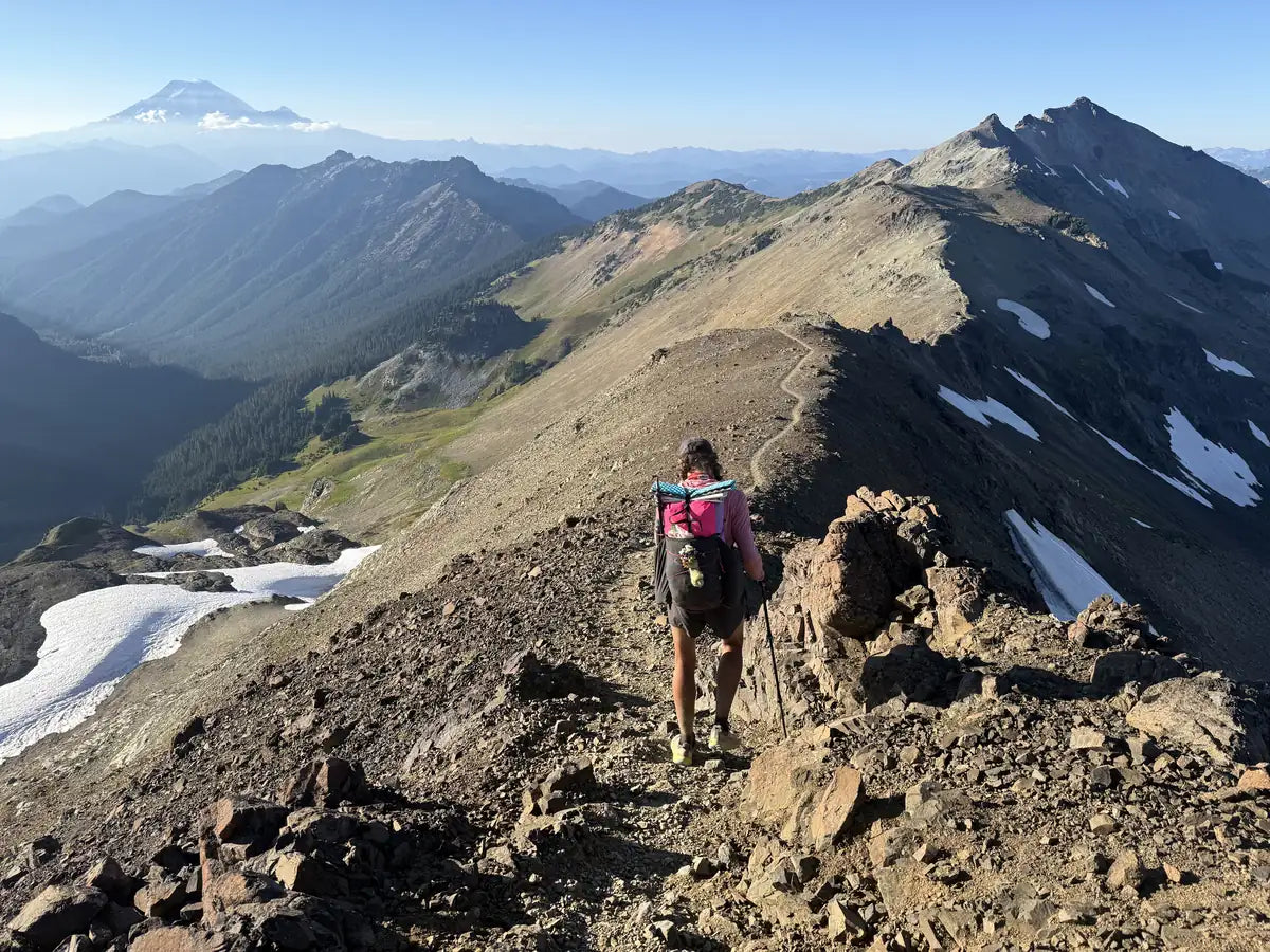 Jocelyn Smith hiking a rocky mountain ridge above forested valleys and patches of snow