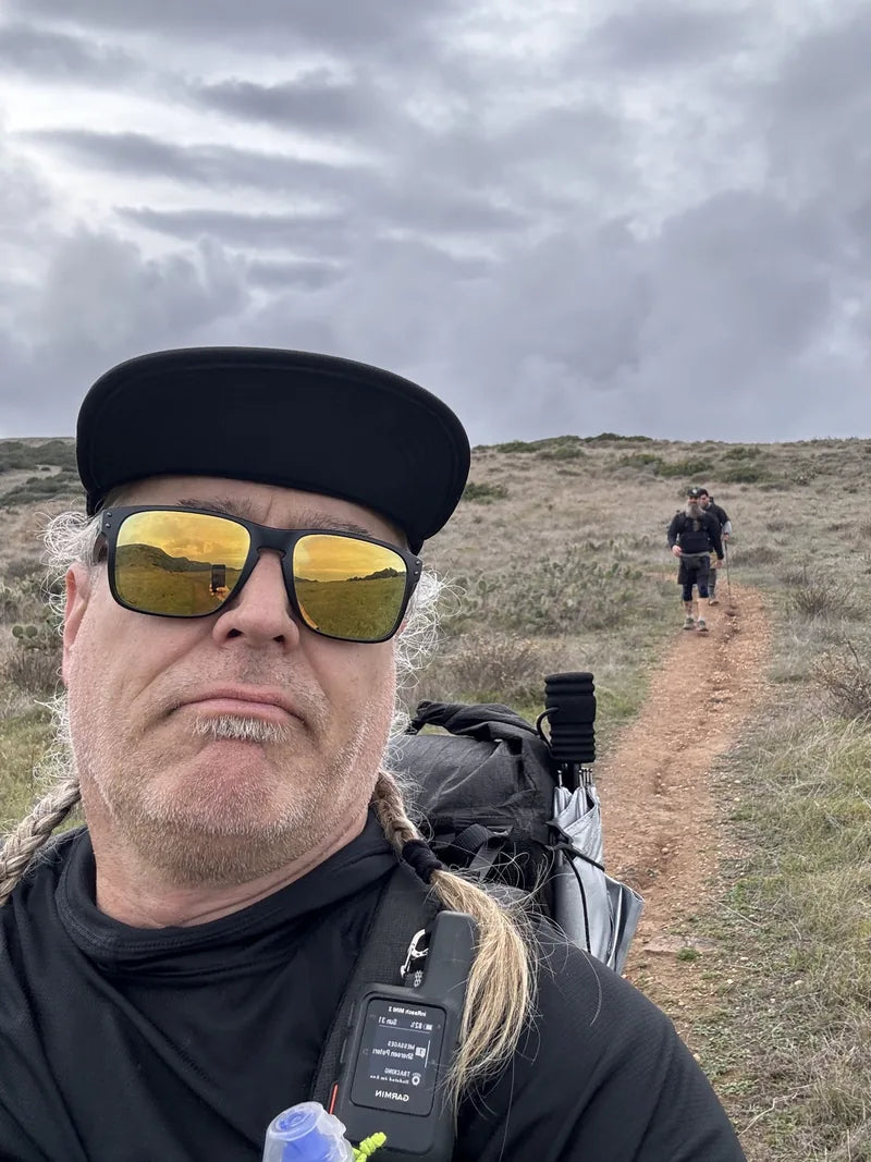 A backpacker taking a trail selfie with two fellow hikers behind him